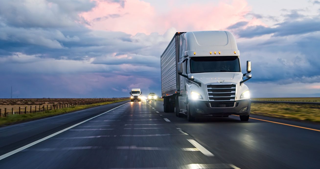 White-semi-truck-driving-on-highway-with-headlights-on-at-dusk