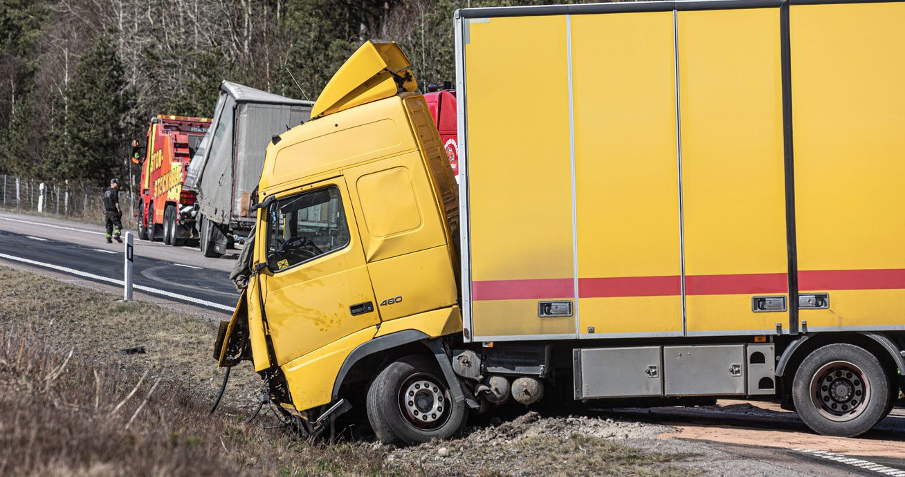 Yellow-box-truck-crashed-off-roadside-with-tow-truck-assistance-in-background